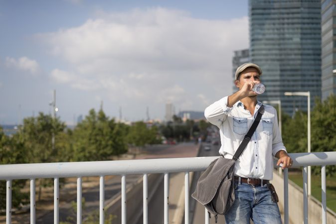 Confident male in denim with city in background drinking from water bottle, copy space