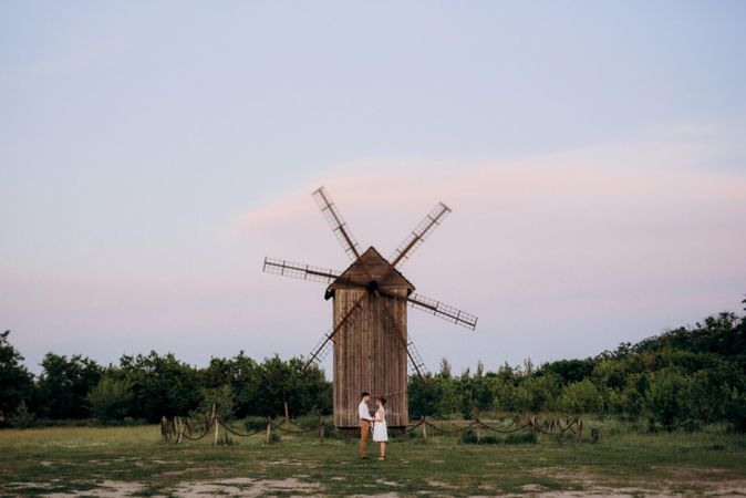 Man and woman holding hands and standing beside wooden windmill on green grass field