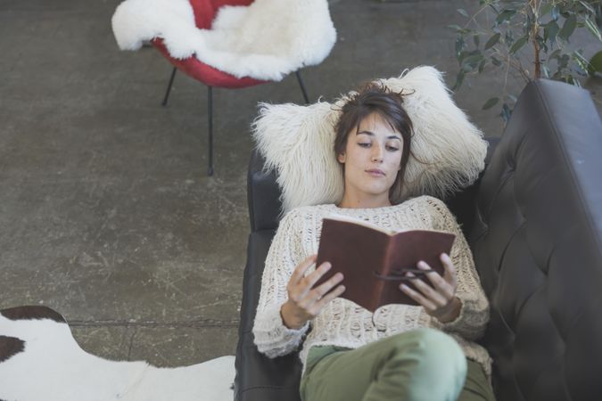 Looking down at female lying back reading book on leather sofa