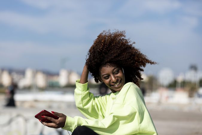 Smiling female with curly hair in bright green shirt standing listening to music on pier