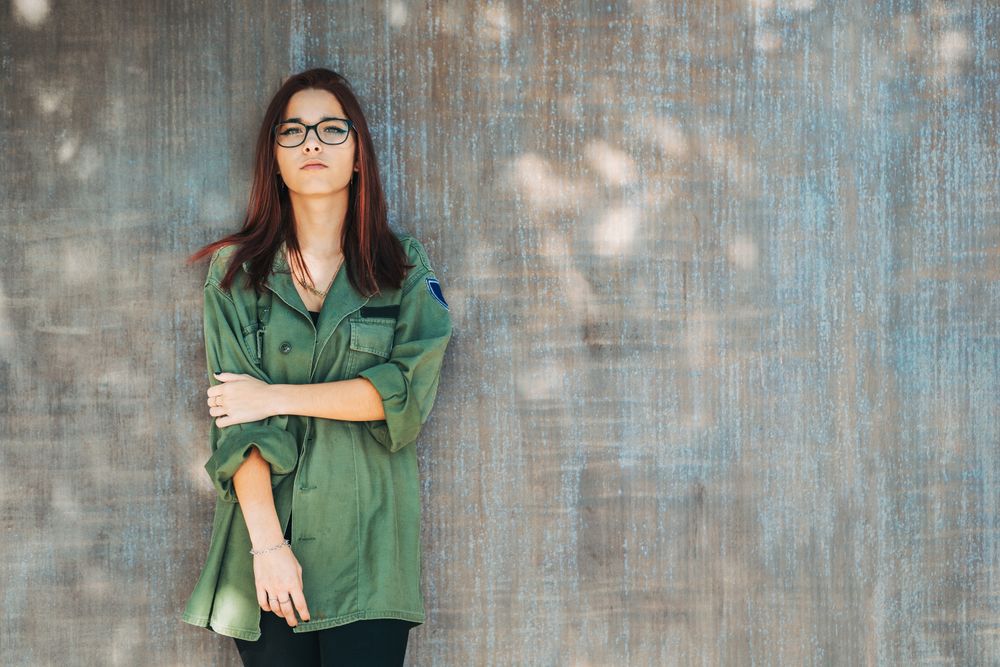 Stylish confident bright woman in glasses leaning on wall and looking at camera