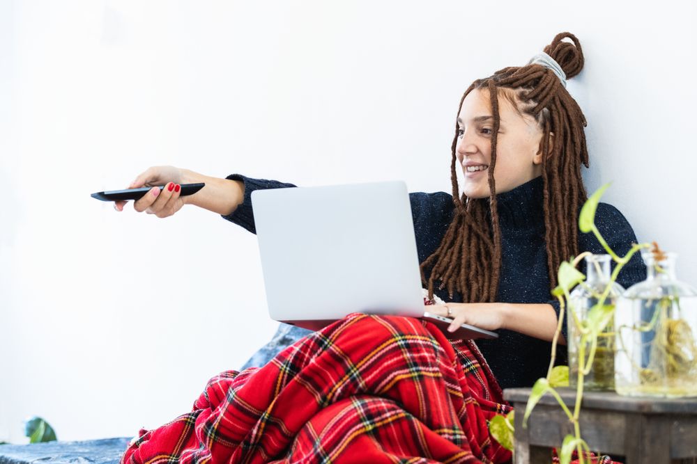 Woman with dreadlocks working with a laptop at home and with a remote control in hand