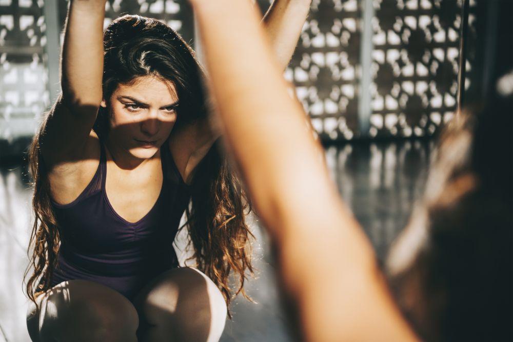 Concentrated brunette in ballet class warming up while looking at mirror in sunlight.