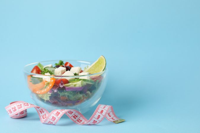 Glass bowl with fresh salad and measuring tape on blue background, space for text