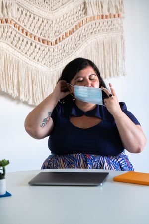 Woman sitting in a modern office with her laptop, adjusting her face mask