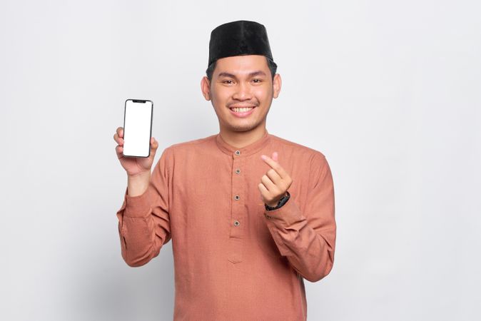 Muslim man in kufi hat smiling with mobile phone making heart sign with fingers
