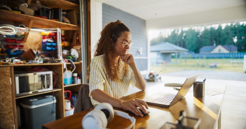 Happy Hispanic Female Engineering Student Sitting At Desk And Using Laptop Computer In Opened Garage.