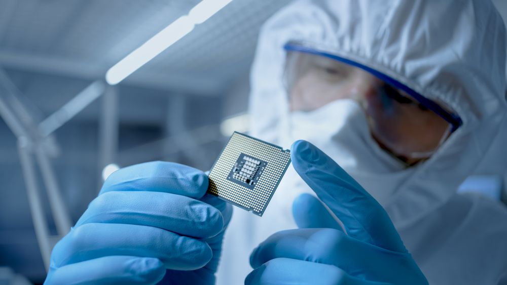In Ultra Modern Electronic Manufacturing Factory Design Engineer in Sterile Coverall Holds Microchip with Gloves and Examines it.