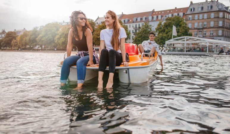 Two young female friends sitting in front of boat with their feet in the water