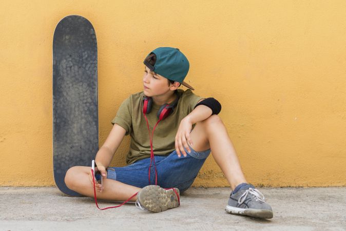 Front view of a teenager sitting on ground leaning on a yellow wall while holding a mobile phone