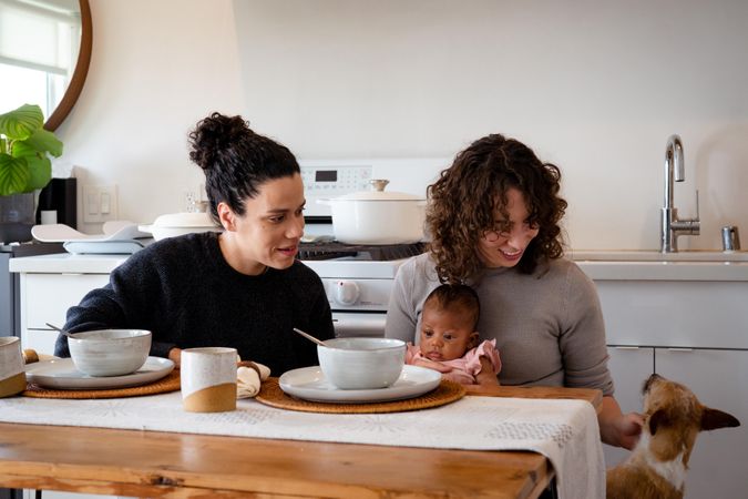 Two women with newborn baby petting their dog at their dining table