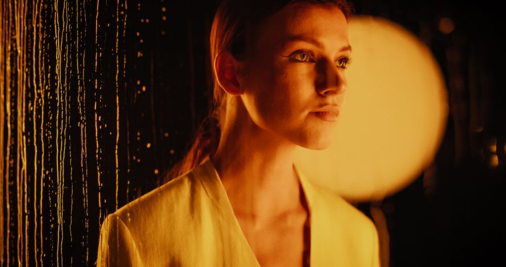 Beauty Footage in a Dark Space with Rain Drops on Glass: Young Female Poses with Confidence, Looking Straight into Camera, Standing Behind a Wet Glass Wall with Small Waterfalls