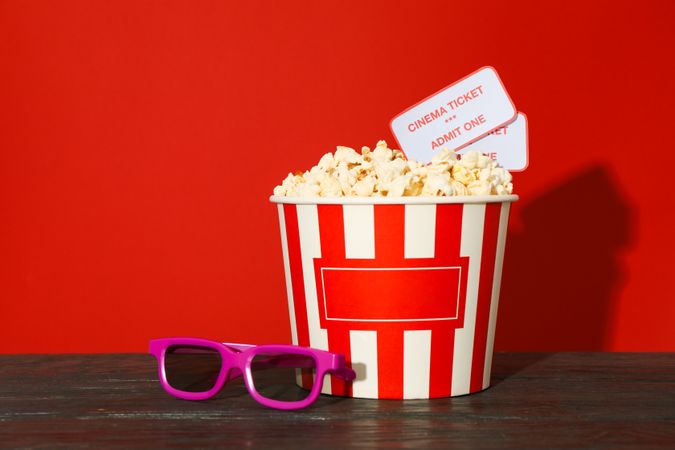 3d glasses, striped bucket with popcorn and tickets against red background