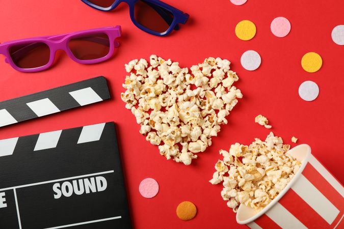 Heart laid out from popcorn, clapperboard, 3d glasses and bucket on red background