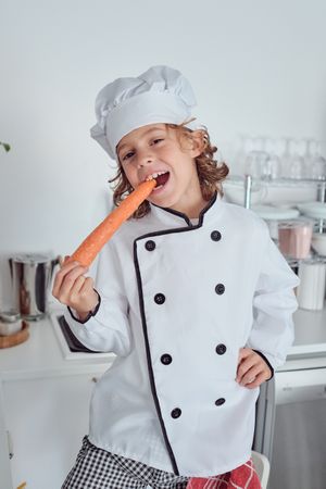 Cheerful boy in chef uniform eating carrot