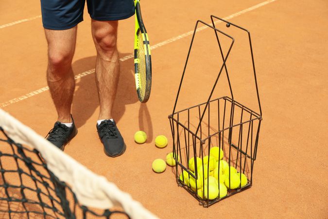 Man hold racket on clay court with basket of tennis balls