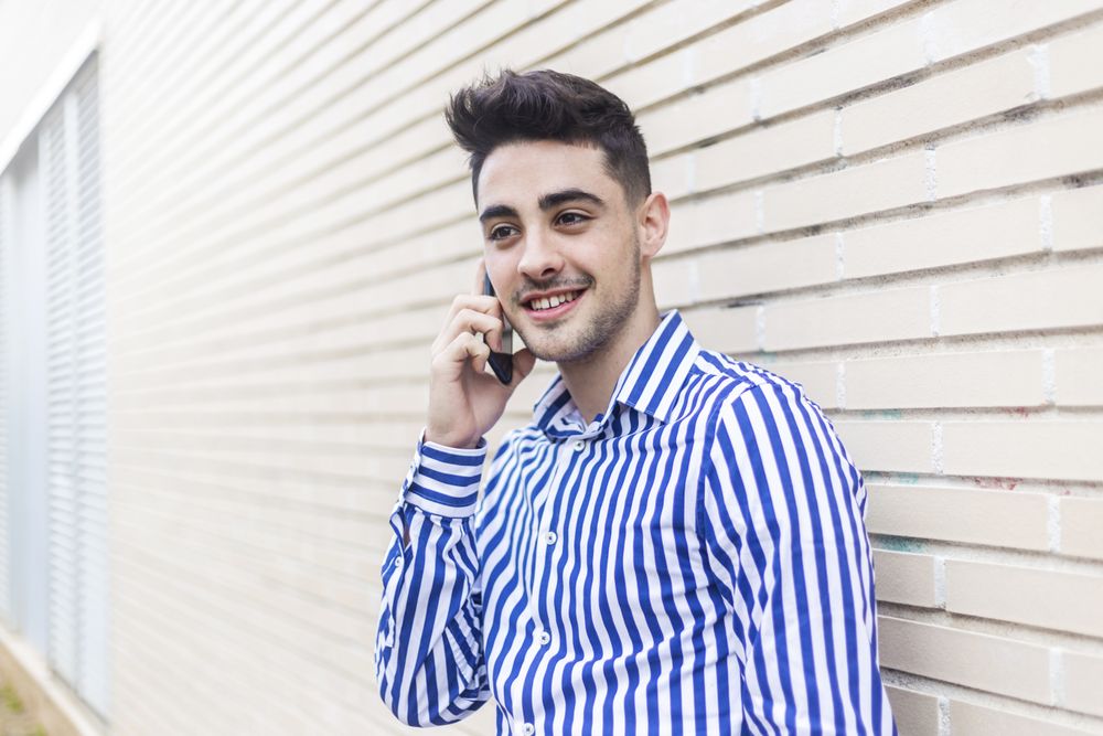 Smiling man leaning on house wall talking on his smartphone and looking into the distance