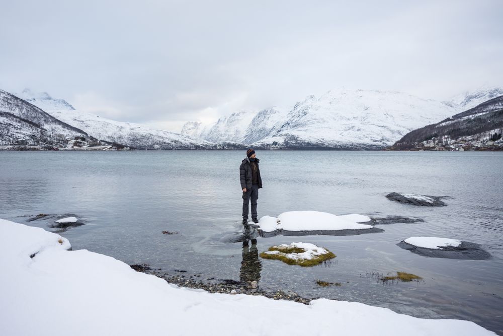 Solitary tourist on fjord shore against snowy hills in cold overcast weather
