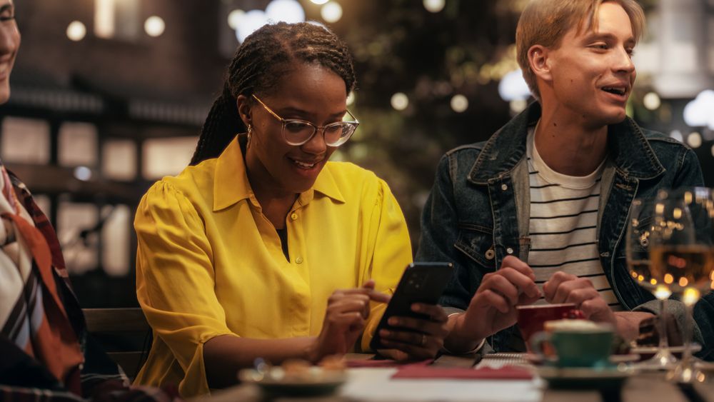 Young Black Female Focused On Smartphone, Not Paying Attention to a Group of Friends Sitting in a Cafe Terrace Around Her