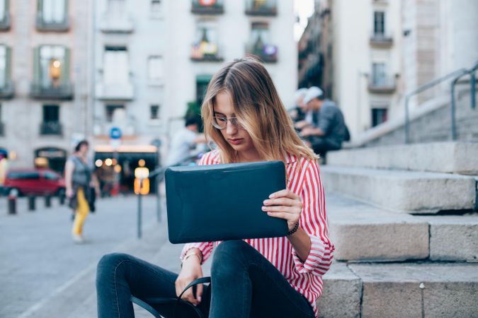 Chic woman sitting on outdoor steps in European city with laptop