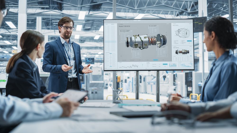 Electronics Factory Office Meeting: Male Project Manager Presenting Innovative Engine On TV.