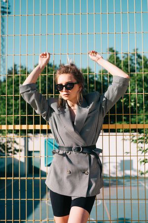 Stylish woman in sunglasses raising hands and leaning on lattice