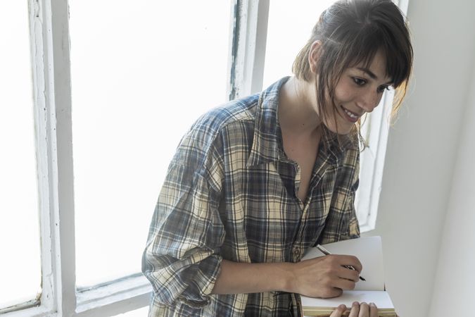 Female in flannel shirt writing in notebook leaning on window