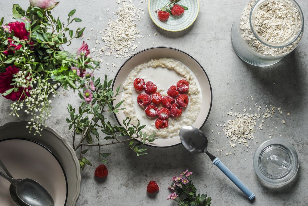 Bowl with porridge and heart shaped raspberries at grey concrete kitchen table with glass jar, branches, bowls and spoon. Preparing healthy vegan breakfast at home. Top view.