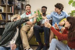 Diverse group of young friends toasting with beer bottles on a sofa at home.