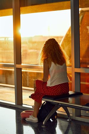 Stylish cheerful woman sitting on metal bench in glass hallway of airport in Texas