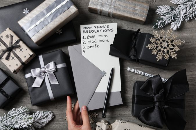 Envelope in female hand, paper with list and gift boxes on gray wooden background, top view