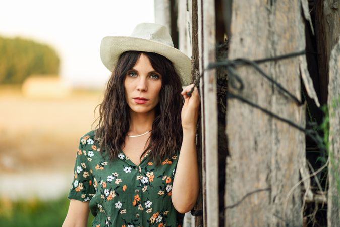 Serious female brunette in straw hat leaning against wooden wall in field