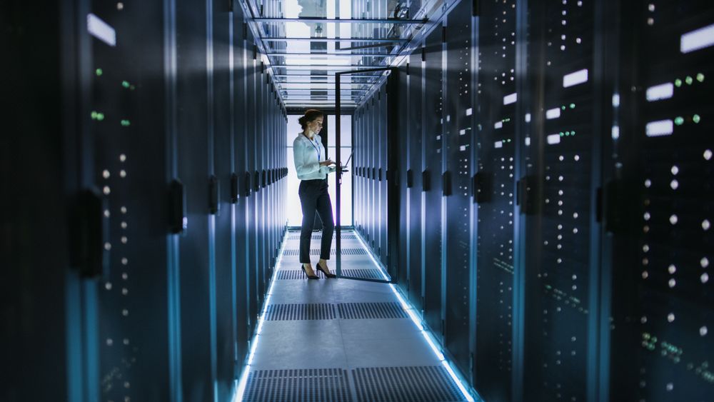 Female Server Technician Stands next to Cabinet in Data Center Corridor with Rows of Rack Servers.