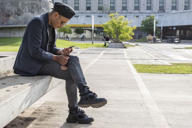 Young Latino gay man wearing a fashionable hat and sunglasses, texting on his smartphone while sitting on a beach outdoors.
