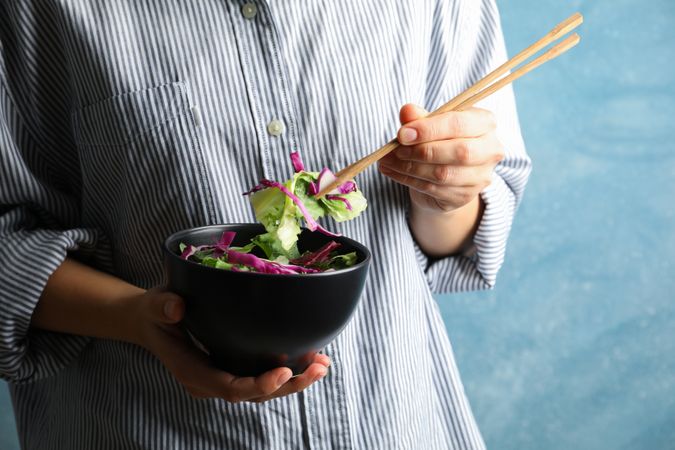 Young woman eat salad with chopsticks on blue background, space for text