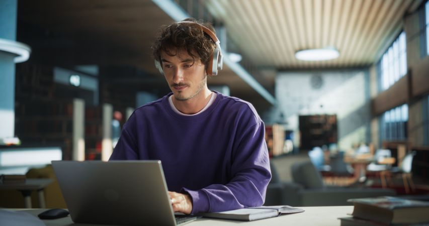 Male Student Writing an Essay on Laptop Computer, Thinking About the Topic, Brainstorming, Finding Solutions. Young Stylish Man Studying in a Public Library, Listening to Lecture in Headphones