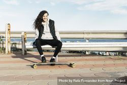 Teenage girl sitting on the bench smiling with her feet on the longboard in front of the sea 5Q28em