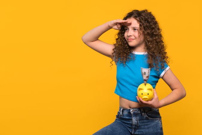 Girl holding piggy bank and saluting, symbolizing future financial planning.