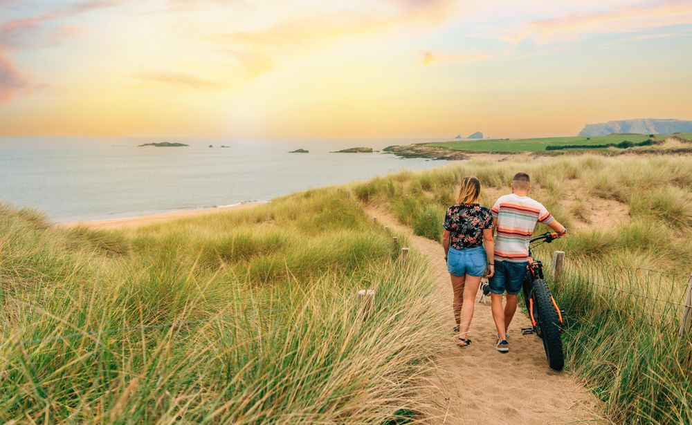 Back of couple walking with bicycle on sandy path towards the ocean