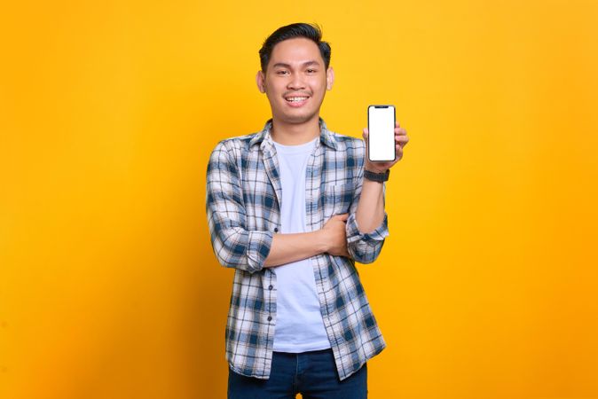 Happy Asian male showing blank screen of smart phone with arms crossed in studio shoot
