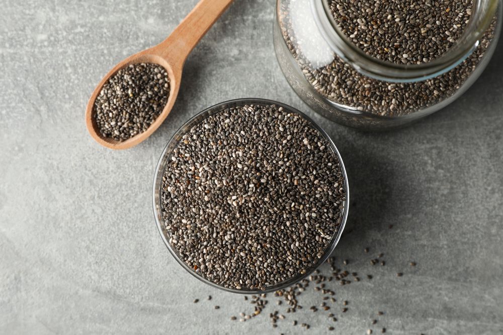 Glass bowl, jar and wood spoon with chia on grey background, top view