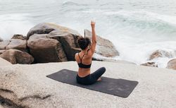 Female sitting next to the ocean sitting crossed legged stretching one arm up