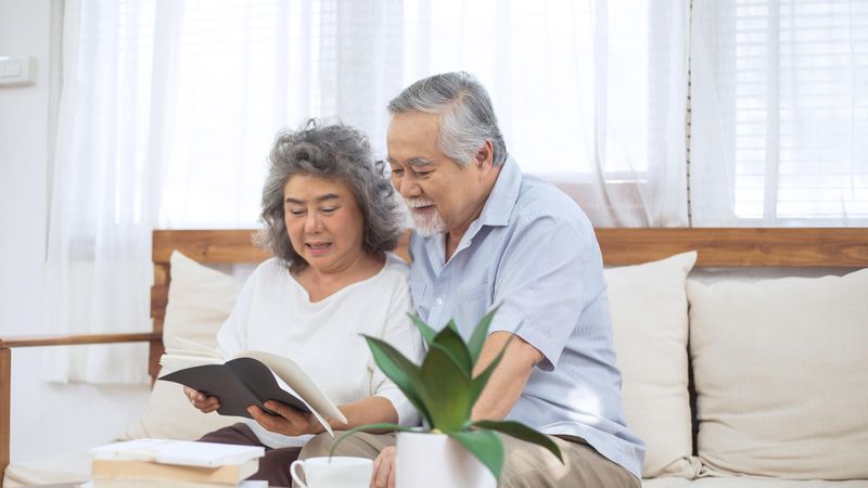 Happy mature Asian couple reading book together on couch in living room