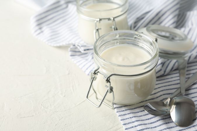 Glass jars with sour cream yogurt, spoons and towel on cement background