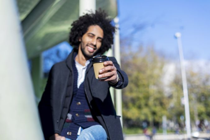 Happy Black man in jacket holding out warm beverage to camera