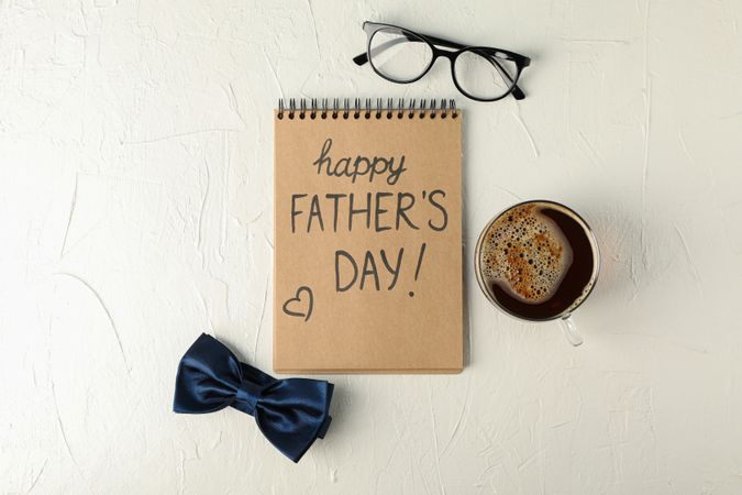 Notebook with inscription happy fathers day, blue bow tie, cup of coffee and glasses on light background, space for text and top view