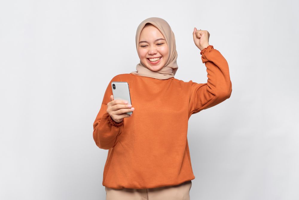 Muslim woman winning something in headscarf and orange shirt holding phone