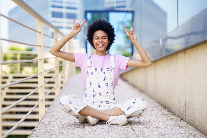 Woman in floral overalls sitting cross legged with hands up