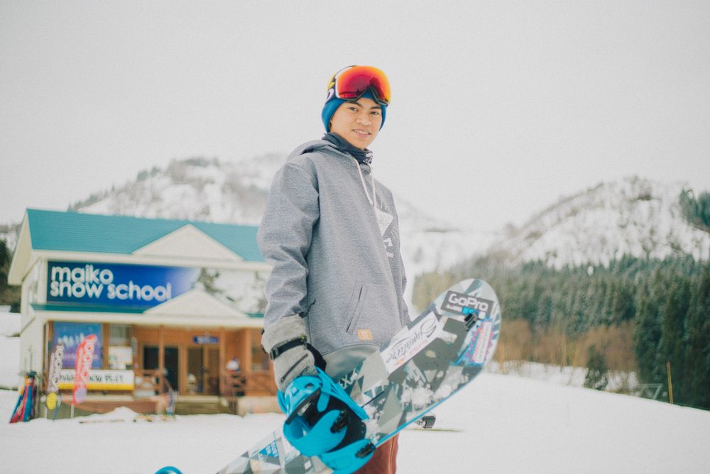 Man in gray jacket holding a snowboard near snow-covered mountains