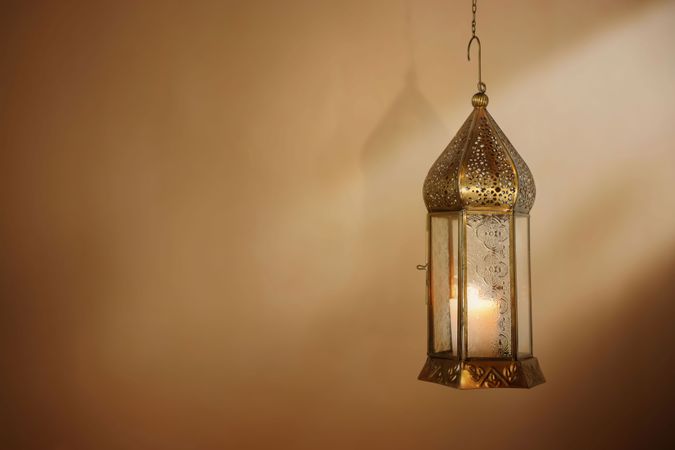 Hanginng decorative brass Moroccan lantern glowing at night. Blurred golden background with light beams. Ramadan Kareem, Eid ul Fitr muslim holiday still life. Festive celebration concept, banner.